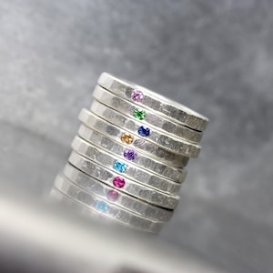 May include: A stack of seven silver rings, each featuring a single gemstone. The gemstones are pink, blue, purple, orange, green, blue, and red.
