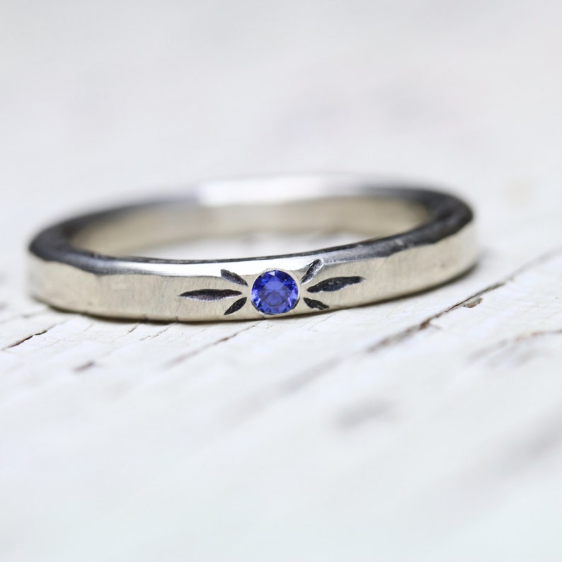 May include: A silver band ring with a small blue sapphire gemstone set in the center. The band has a textured surface and a simple design.