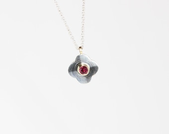 Puede incluir: Collar plateado con un colgante de trébol de cuatro hojas. El colgante tiene un acabado gris texturizado con un círculo interior dorado. Una pequeña gema roja redonda está engastada en el centro del colgante.