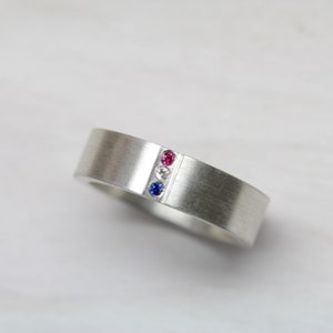 May include: A silver band ring with a vertical row of three gemstones: a red ruby, a white diamond, and a blue sapphire.