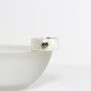 May include: A silver ring with a textured band and a small heart-shaped blue gemstone set in gold.