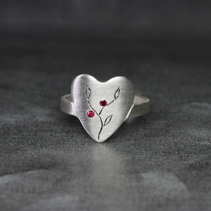 May include: A silver heart-shaped ring with a delicate branch design and two small red gemstones.