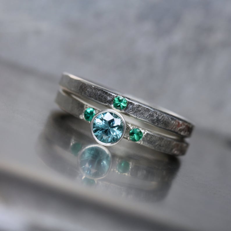 May include: Two silver rings, one stacked on top of the other. The top ring has three small green gemstones. The bottom ring has a larger, round, light blue gemstone in the center, flanked by two smaller green gemstones.