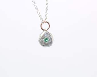 Puede incluir: Un collar de plata con un pequeño colgante de forma irregular que presenta una piedra preciosa verde en el centro.