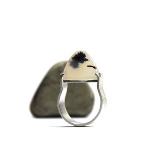 May include: A silver ring with a white agate stone set in the center. The stone has black dendritic patterns. The ring is sitting on a gray rock.