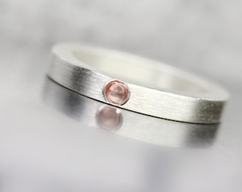 May include: A silver band ring with a single, round, light pink gemstone set in the center.