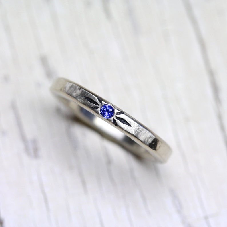 May include: A silver band ring with a small blue sapphire gemstone set in the center. The band has a hammered texture and a simple design.