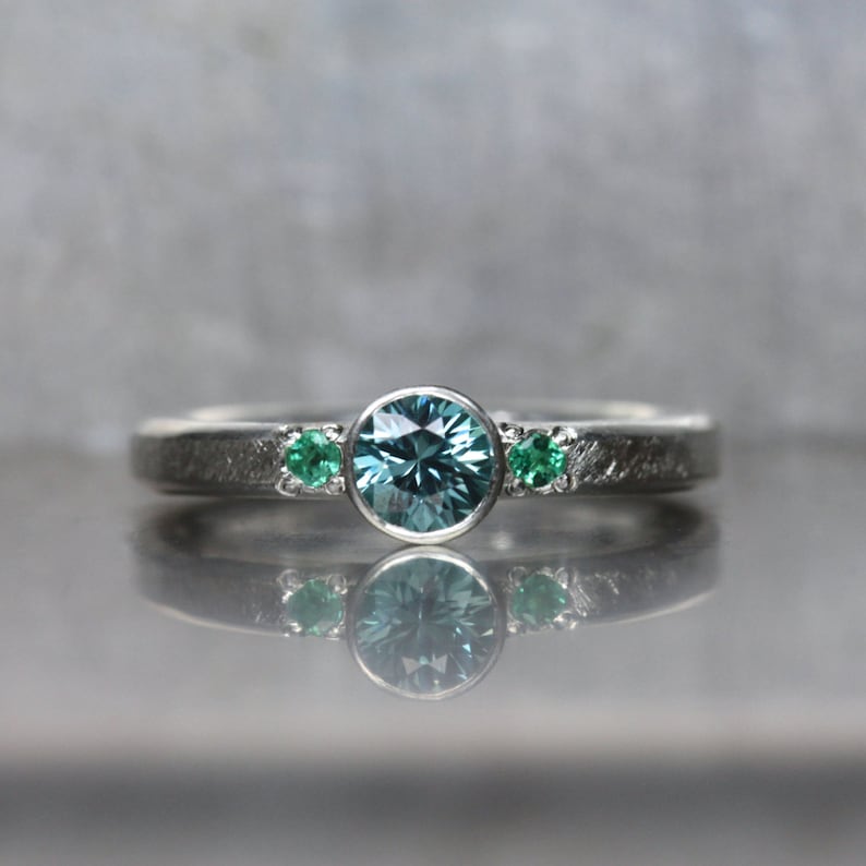 May include: A silver ring with a large, round, light blue gemstone in the center, flanked by two smaller green gemstones. The band has a textured finish, and the ring is displayed on a reflective surface.