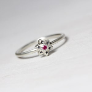May include: A silver ring with a red gemstone in the center of a star-shaped design.