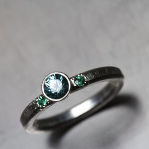 May include: A silver ring with a dark, textured band. The ring features a large, round, teal-colored gemstone in the center, flanked by two smaller, emerald-green gemstones. The gemstones are set in a minimalist style.