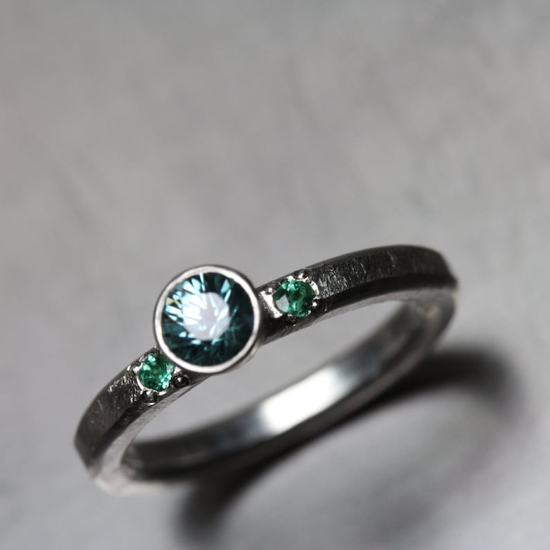 May include: A silver ring with a dark, textured band. The ring features a large, round, teal-colored gemstone in the center, flanked by two smaller, emerald-green gemstones. The gemstones are set in a minimalist style.