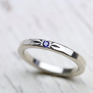 May include: A silver band ring with a small blue gemstone set in the center. The band has a textured design with a starburst pattern.