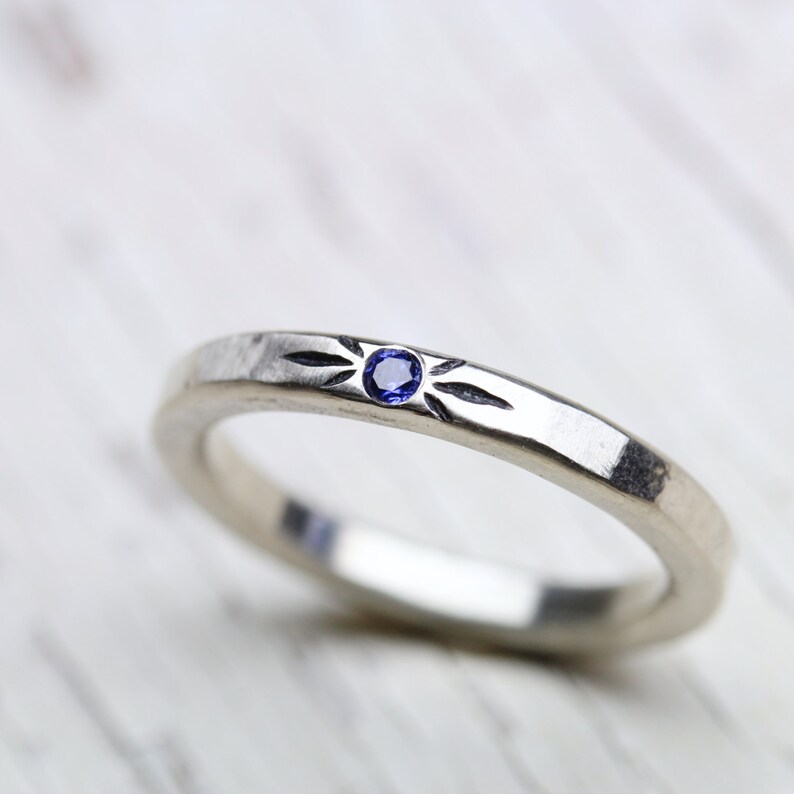 May include: A silver band ring with a small blue gemstone set in the center. The band has a textured design with a starburst pattern.