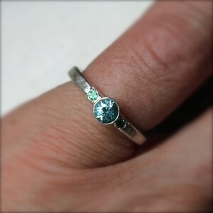 May include: A silver ring with a large, round, light blue gemstone in the center, flanked by two smaller green gemstones. The ring is set on a finger, showcasing the delicate design and color contrast.