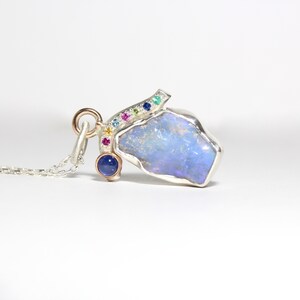May include: A silver necklace with a pendant featuring a large, blue-grey opal stone. The pendant is accented with a row of colorful gemstones, including pink, yellow, green, and blue.
