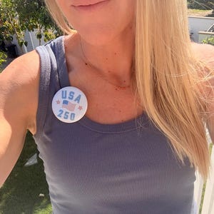 Puede incluir: Primer plano de una persona con una camiseta sin mangas azul acanalada. Un botón redondo prendido en la parte superior muestra el texto "USA 250" con una pequeña bandera estadounidense. La persona tiene cabello largo y rubio y un delicado collar.