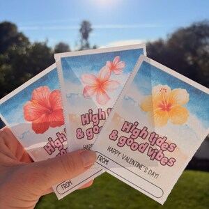 May include: Three Valentine's Day cards with watercolor floral designs and the text "High tides & good vibes". Each card features a different flower: red hibiscus, pink plumeria, and yellow hibiscus. The cards have a blue watercolor background.