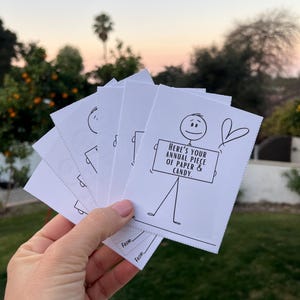May include: A set of five white cards with black ink drawings of a stick figure holding a sign that says "Here's your annual piece of paper & candy." The cards have the words "From:" written on them.