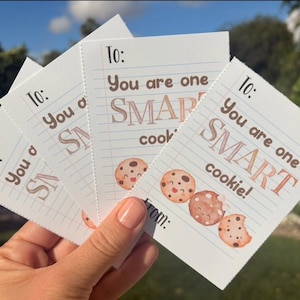 May include: A stack of white cards with a lined paper design. Each card has the text "To: You are one SMART cookie!" in brown and a watercolor illustration of smiling cookies. The cards are held in a hand.