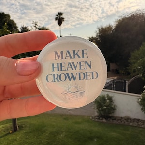 May include: A round, white button with the text "MAKE HEAVEN CROWDED" in blue script. Below the text is a graphic of a sun over water. The button is held in front of a green lawn and a cloudy sky.