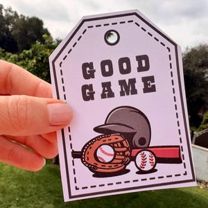 May include: A light purple tag with the words "GOOD GAME" in bold, black letters. Below the text is a baseball helmet, glove, ball, and a red bat. The tag has a black border and a hole at the top.