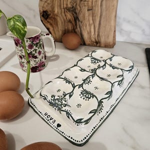 Può includere: Un portauova in ceramica bianca con un motivo floreale verde scuro. Il portauova ha spazio per sei uova e la parola "uova" è scritta sul lato. Sono visibili anche una brocca floreale e uova marroni.