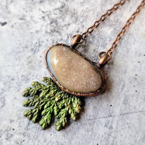 May include: A copper-toned necklace featuring a druzy stone pendant with a light brown, sparkling surface. The pendant is set in a copper frame and hangs from a delicate chain. A green, leaf-like embellishment rests beneath the pendant.
