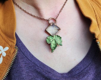 May include: A copper-toned necklace featuring a diamond-shaped pendant with a clear center and a green leaf charm. The necklace is worn against a purple shirt and a mustard-colored jacket.