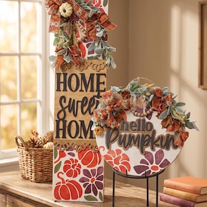 Peut inclure: Un panneau rectangulaire en bois avec le texte HOME sweet HOME et un panneau rond avec le texte hello Pumpkin. Les deux pr&eacute;sentent des motifs de citrouilles et de fleurs orange et violettes, orn&eacute;s de n&oelig;uds en tissu, de feuillage et de citrouilles.