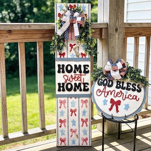 May include: A wooden sign with the words "HOME SWEET HOME" and "GOD BLESS AMERICA" in red, white, and blue. The signs are decorated with bows, stars, and a wreath, perfect for patriotic home decor.