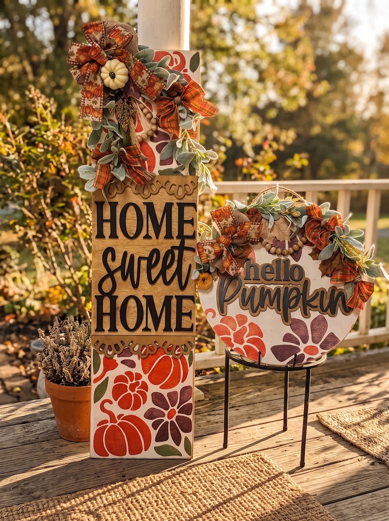 Peut inclure: Un panneau en bois rectangulaire haut avec le texte HOME sweet HOME et un panneau rond avec le texte hello Pumpkin. Les deux panneaux pr&eacute;sentent des citrouilles peintes orange et violettes, des feuilles vertes, des n&oelig;uds en tissu &eacute;cossais.