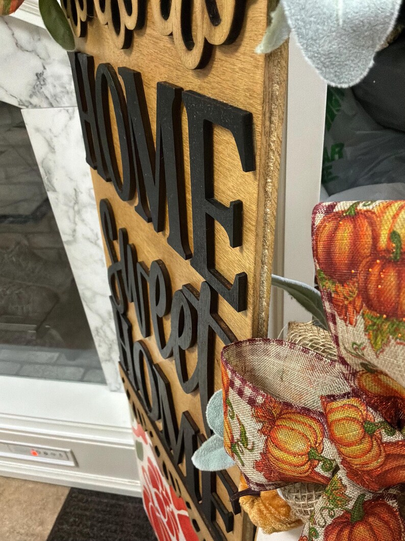 Peut inclure: Un panneau vertical en bois pr&eacute;sentant le texte d&eacute;coup&eacute; noir Home Sweet Home superpos&eacute; sur un fond en bois clair. Le panneau est d&eacute;cor&eacute; d'un ruban en toile de jute &agrave; motifs de citrouilles orange et de feuilles vertes fix&eacute; sur le c&ocirc;t&eacute;.