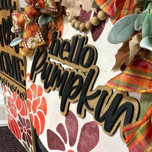 Peut inclure: Une plaque de porte ronde en bois avec le texte noir en relief hello pumpkin, orn&eacute;e de motifs floraux orange et rouges, d'un ruban &agrave; carreaux, de toile de jute, de perles en bois et de feuillage vert. Une plaque rectangulaire est visible &agrave; gauche.