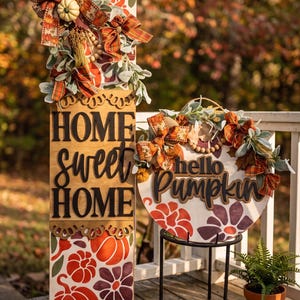 Peut inclure: Un panneau en bois rectangulaire haut avec le texte HOME sweet HOME et un panneau rond en bois avec le texte hello Pumpkin. Les deux panneaux pr&eacute;sentent des motifs de citrouilles et de fleurs orange et violettes, des n&oelig;uds en toile de jute.