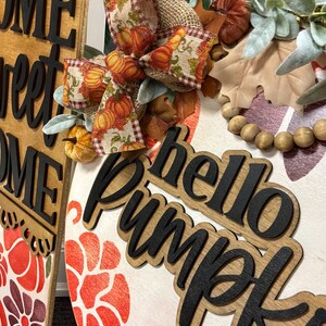 Peut inclure: Une plaque de porte ronde en bois avec le texte hello Pumpkin en lettres 3D noires. La plaque pr&eacute;sente un n&oelig;ud en ruban de toile de jute &agrave; motifs de citrouilles, des feuilles vertes artificielles, des perles en bois et des motifs floraux peints.
