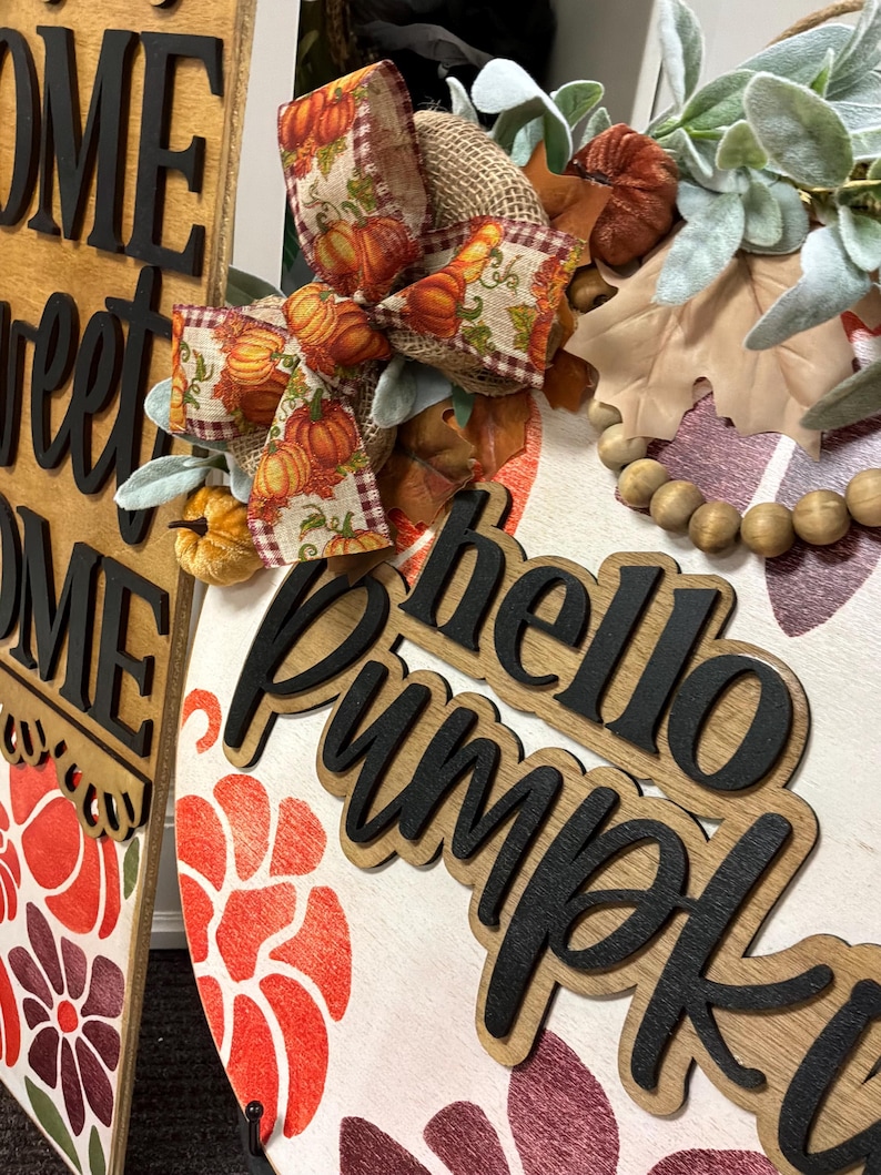 Peut inclure: Une plaque de porte ronde en bois avec le texte hello Pumpkin en lettres 3D noires. La plaque pr&eacute;sente un n&oelig;ud en ruban de toile de jute &agrave; motifs de citrouilles, des feuilles vertes artificielles, des perles en bois et des motifs floraux peints.