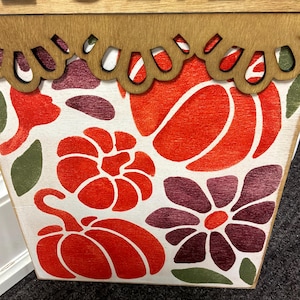 Peut inclure: Un panneau en bois rectangulaire avec un fond blanc orn&eacute; d'un motif imprim&eacute; de citrouilles orange, de fleurs violet fonc&eacute; et de feuilles vertes. Le bord sup&eacute;rieur pr&eacute;sente une bordure en bois festonn&eacute;e et des lettres noires.
