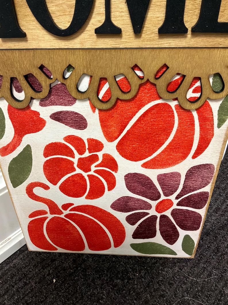 Peut inclure: Un panneau en bois rectangulaire avec un fond blanc orn&eacute; d'un motif imprim&eacute; de citrouilles orange, de fleurs violet fonc&eacute; et de feuilles vertes. Le bord sup&eacute;rieur pr&eacute;sente une bordure en bois festonn&eacute;e et des lettres noires.