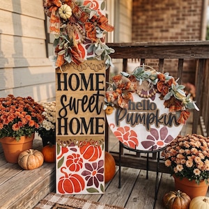 Peut inclure: Un grand panneau en bois rectangulaire avec le texte HOME sweet HOME et un panneau rond en bois avec le texte hello Pumpkin. Les deux panneaux pr&eacute;sentent des citrouilles orange et violettes, des motifs floraux et des n&oelig;uds d&eacute;coratifs.