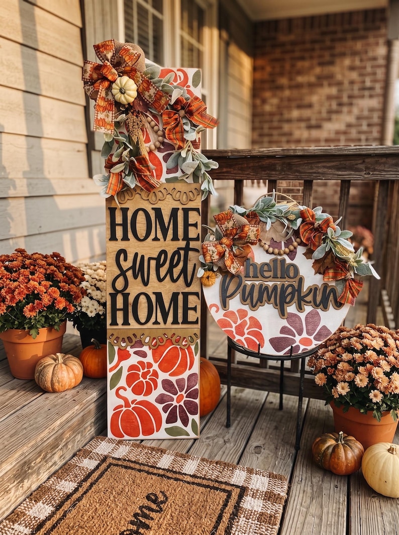 Peut inclure: Un grand panneau en bois rectangulaire avec le texte HOME sweet HOME et un panneau rond en bois avec le texte hello Pumpkin. Les deux panneaux pr&eacute;sentent des citrouilles orange et violettes, des motifs floraux et des n&oelig;uds d&eacute;coratifs.