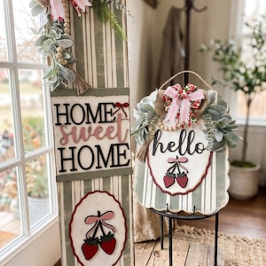 May include: Two decorative wooden signs with a farmhouse theme. One sign reads "HOME SWEET HOME" and features a strawberry design. The other sign says "hello" and also has a strawberry design. Both signs are decorated with bows and greenery.