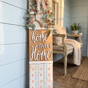 May include: A vertical wooden sign with the words "home sweet home" in white script. The sign is decorated with a floral wreath, a burlap bow, and a patterned design in shades of blue, peach, and white. The sign is leaning against a light blue wall.