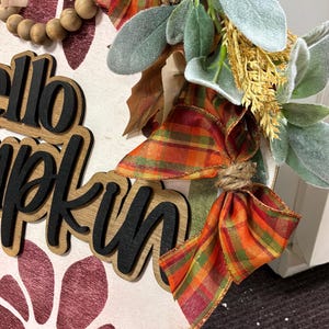 Peut inclure: Une plaque de porte ronde en bois avec le texte "hello pumpkin" en lettres noires 3D. La plaque est d&eacute;cor&eacute;e d'un ruban &agrave; carreaux orange, vert et jaune, de perles en bois, de feuilles vert sauge artificielles et d'un motif floral bordeaux.