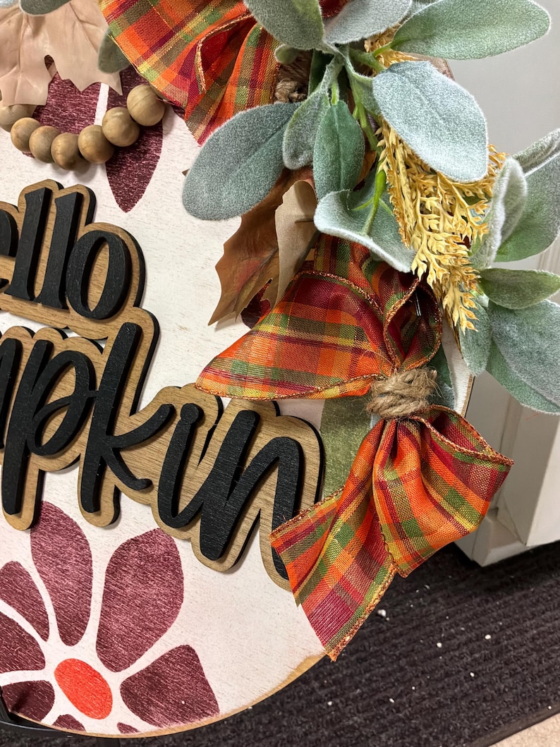 Peut inclure: Une plaque de porte ronde en bois avec le texte "hello pumpkin" en lettres noires 3D. La plaque est d&eacute;cor&eacute;e d'un ruban &agrave; carreaux orange, vert et jaune, de perles en bois, de feuilles vert sauge artificielles et d'un motif floral bordeaux.