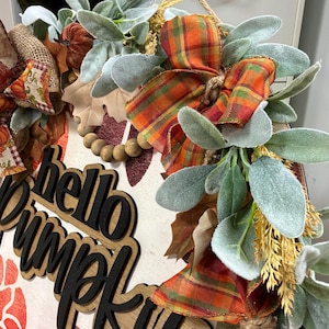Peut inclure: Une plaque de porte ronde en bois avec le texte hello pumpkin en lettres noires 3D. La d&eacute;coration comprend un ruban &agrave; carreaux orange et vert, des feuilles vertes givr&eacute;es artificielles, des perles en bois et des accents en toile de jute.