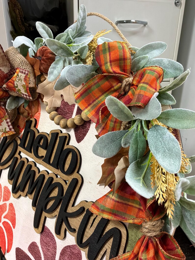 Peut inclure: Une plaque de porte ronde en bois avec le texte hello pumpkin en lettres noires 3D. La d&eacute;coration comprend un ruban &agrave; carreaux orange et vert, des feuilles vertes givr&eacute;es artificielles, des perles en bois et des accents en toile de jute.