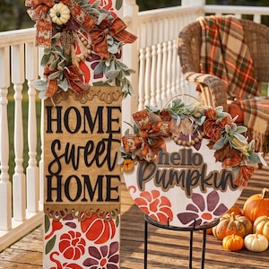 Peut inclure: Un grand panneau rectangulaire en bois avec le texte HOME sweet HOME et un panneau rond en bois avec le texte hello Pumpkin. Les deux panneaux pr&eacute;sentent des motifs de citrouilles et de fleurs orange et violettes, des n&oelig;uds et du feuillage.