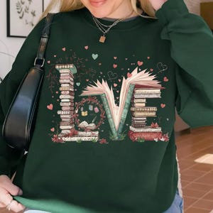 May include: Dark green sweatshirt with the word "LOVE" spelled out with stacks of books and an open book. The design includes hearts and floral accents, creating a literary theme.