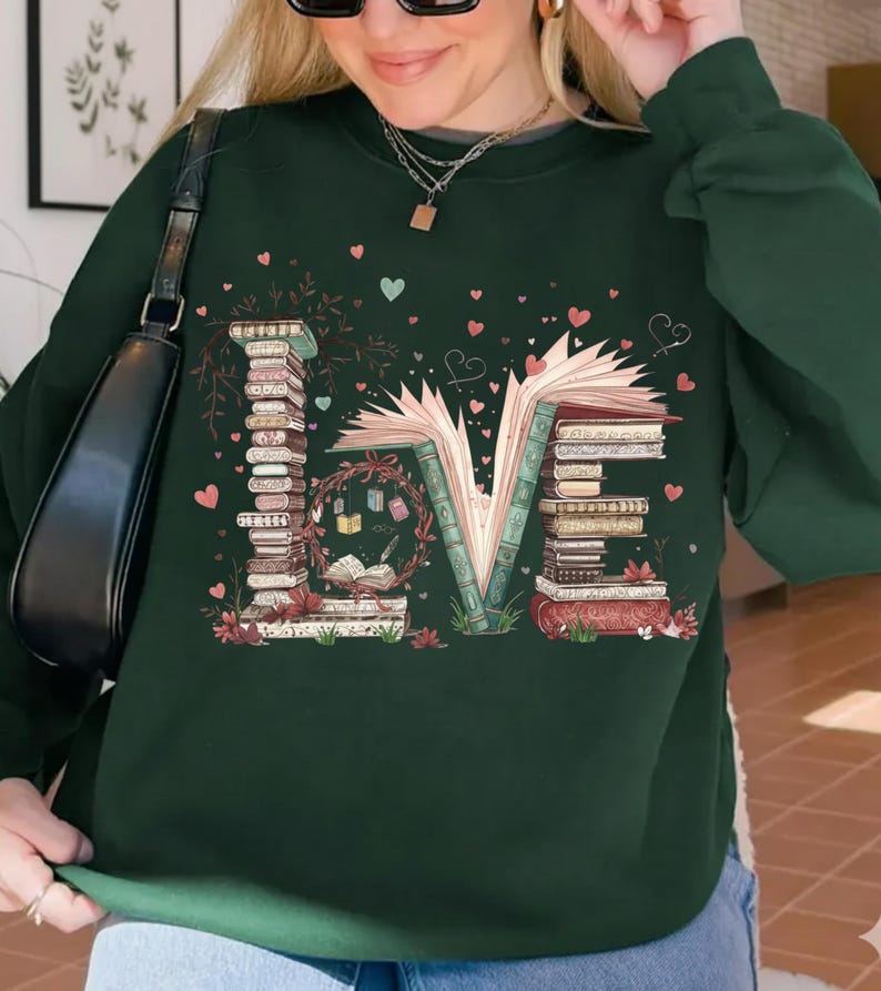 May include: Dark green sweatshirt with the word "LOVE" spelled out with stacks of books and an open book. The design includes hearts and floral accents, creating a literary theme.