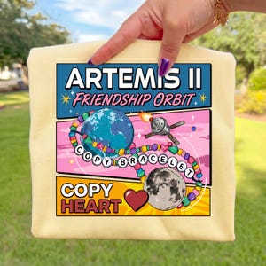 Puede incluir: Camiseta amarillo claro con un diseño gráfico colorido. El diseño incluye el texto "ARTEMIS II FRIENDSHIP ORBIT" sobre una representación de la Tierra, un satélite y la luna. También se incluyen las palabras "COPY BRACELET" y "COPY HEART".